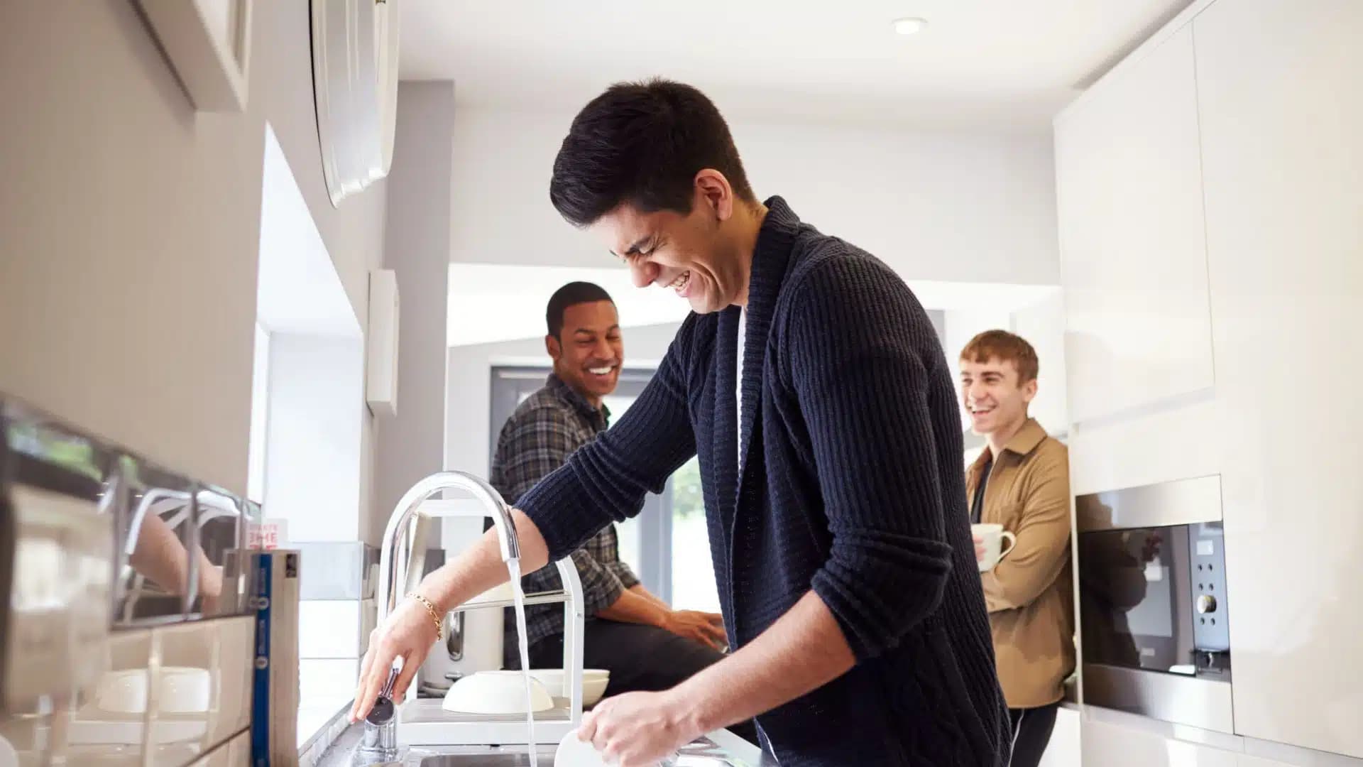 Three men in a kitchen laughing while sink tap is running water